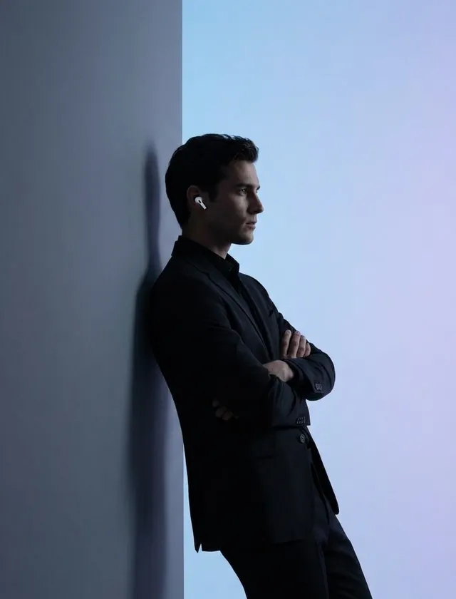 A man in a black suit leans against a wall with blue and gray tones.