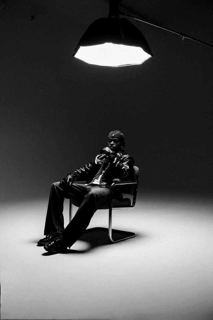 A black and white editorial photograph captures a man seated on a chair, exuding a confident and contemplative mood